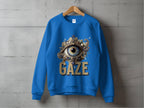 A blue unisex sweatshirt by Garment Graphics hangs on a wooden hanger against a concrete wall. It features a large, detailed eye design with embellishments and the word GAZE boldly in gold letters below.