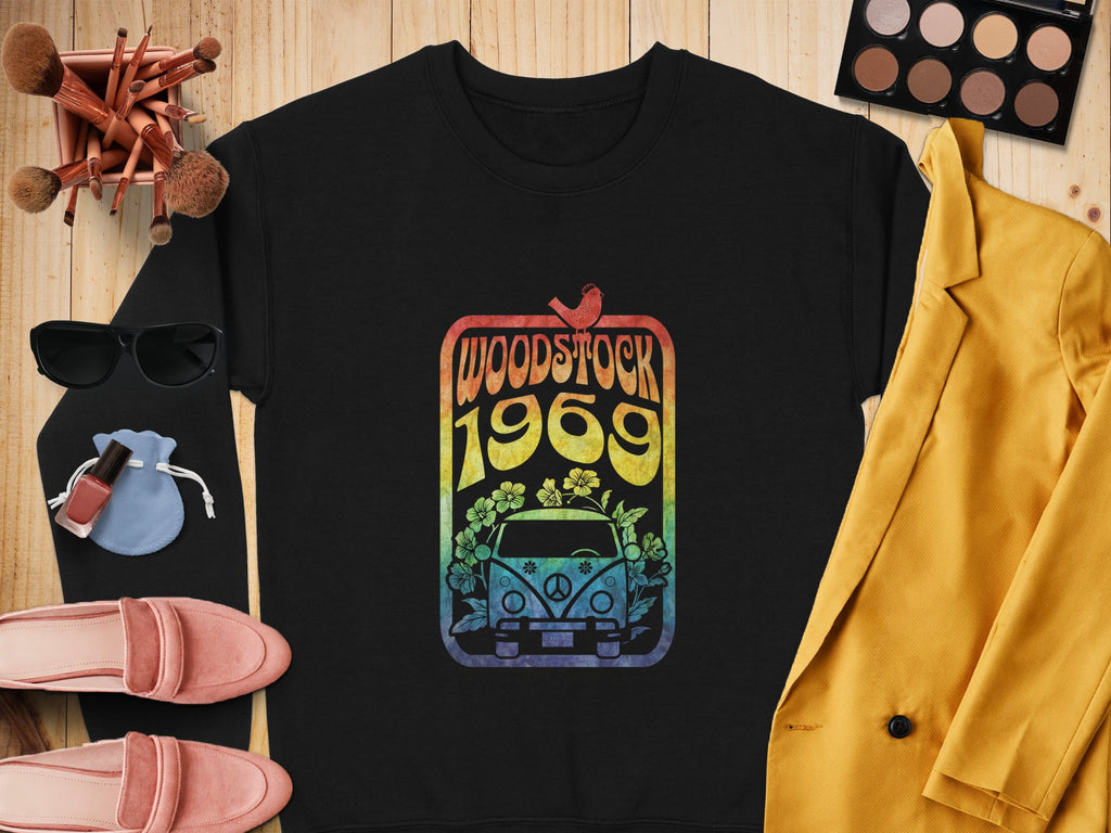 A retro black sweatshirt by Garment Graphics features a colorful Woodstock 1969 design with a car and flowers, surrounded by makeup brushes, palette, sunglasses, coin pouch, nail polish, pink loafers, and mustard yellow blazer.