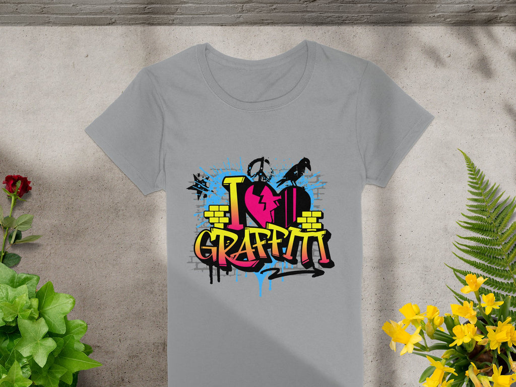 Garment Graphics presents a gray T-shirt with a vibrant street art design, featuring I Love Graffiti text, paint splashes, bricks, and a spray can silhouette. Ideal for urban design enthusiasts, it rests on a concrete surface amidst lush plants.