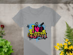 Garment Graphics presents a gray T-shirt with a vibrant street art design, featuring I Love Graffiti text, paint splashes, bricks, and a spray can silhouette. Ideal for urban design enthusiasts, it rests on a concrete surface amidst lush plants.