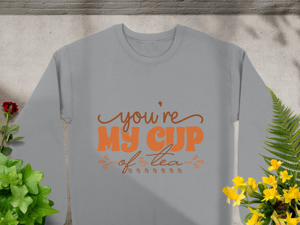 A cozy gray sweatshirt from Garment Graphics, features Youre My Cup of Tea in orange text. Suitable for all with unisex sizing, its pictured laid flat amid green plants and vibrant yellow and red flowers on a gray stone background.