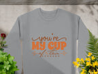 A cozy gray sweatshirt from Garment Graphics, features Youre My Cup of Tea in orange text. Suitable for all with unisex sizing, its pictured laid flat amid green plants and vibrant yellow and red flowers on a gray stone background.