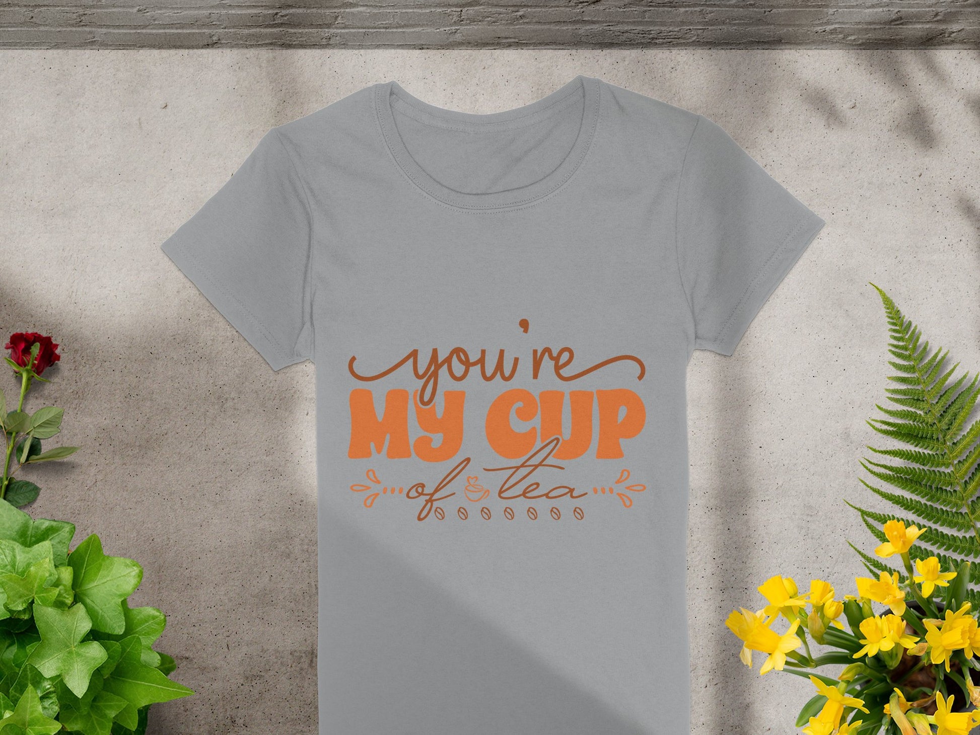 A light grey 100% cotton graphic T-shirt by Garment Graphics features Youre my cup of tea in orange cursive and print font. It rests on concrete amid plants with red and yellow flowers, as leaf shadows dance across the scene.