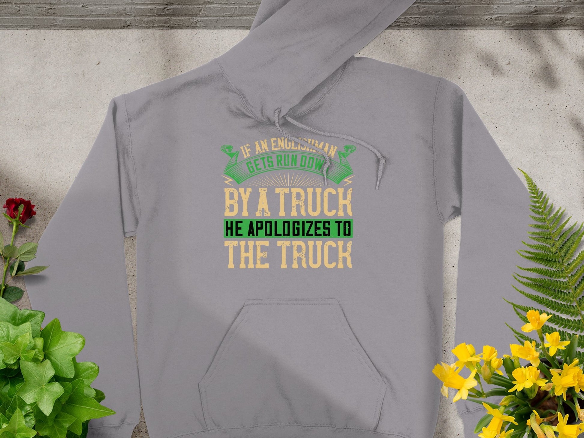 A Garment Graphics gray hoodie lies on a flat surface, surrounded by potted plants. It humorously states, If an Englishman gets run down by a truck, he apologizes to the truck, in playful yellow, green, and black fonts.