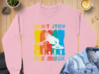 A Garment Graphics pink DJ sweatshirt displays a vivid graphic of a DJ at turntables with vertical stripes and Dont Stop the Music text. Ideal for music enthusiasts, its styled on a wooden table alongside a denim jacket, cappuccino, and small plants.