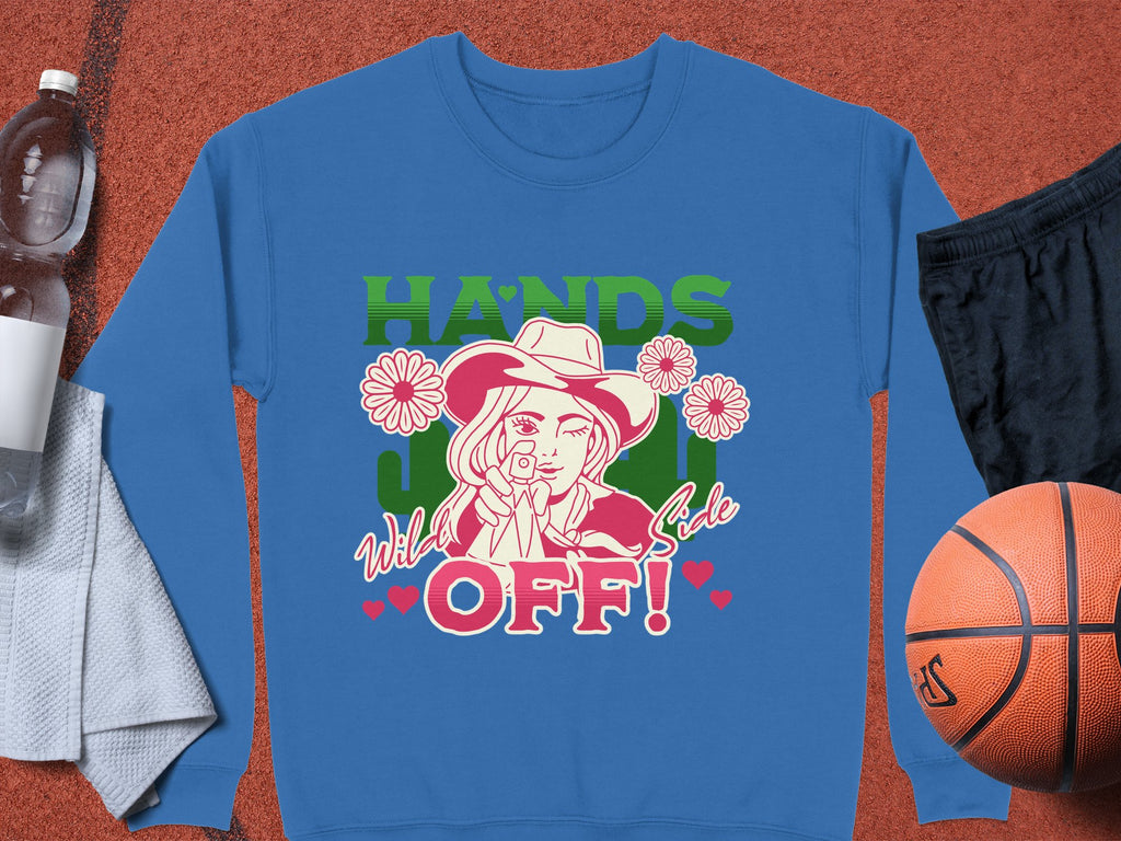 The Garment Graphics Wild Side Sweatshirt, unisex, boasts bold graphics of a hat-wearing figure pointing amid flowers with HANDS OFF! in bright green and red. Its styled on an orange backdrop with a water bottle, towel, black shorts, and a basketball.