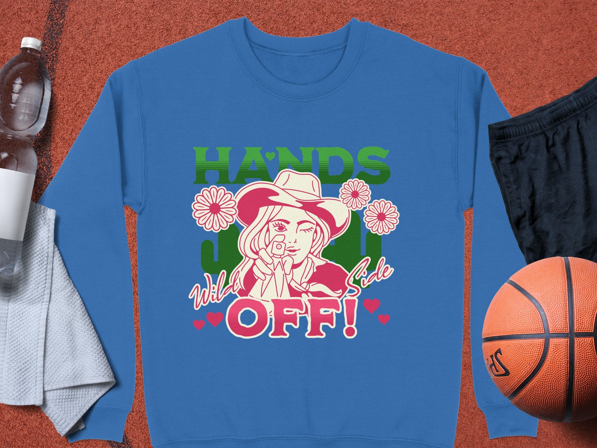 The Garment Graphics Wild Side Sweatshirt, unisex, boasts bold graphics of a hat-wearing figure pointing amid flowers with HANDS OFF! in bright green and red. Its styled on an orange backdrop with a water bottle, towel, black shorts, and a basketball.