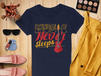 A Garment Graphics navy Music Never Sleeps t-shirt with a guitar graphic lies on a wooden surface, accompanied by a yellow jacket, sunglasses, makeup brushes, an eyeshadow palette, cream slippers, and a perfume bottle.
