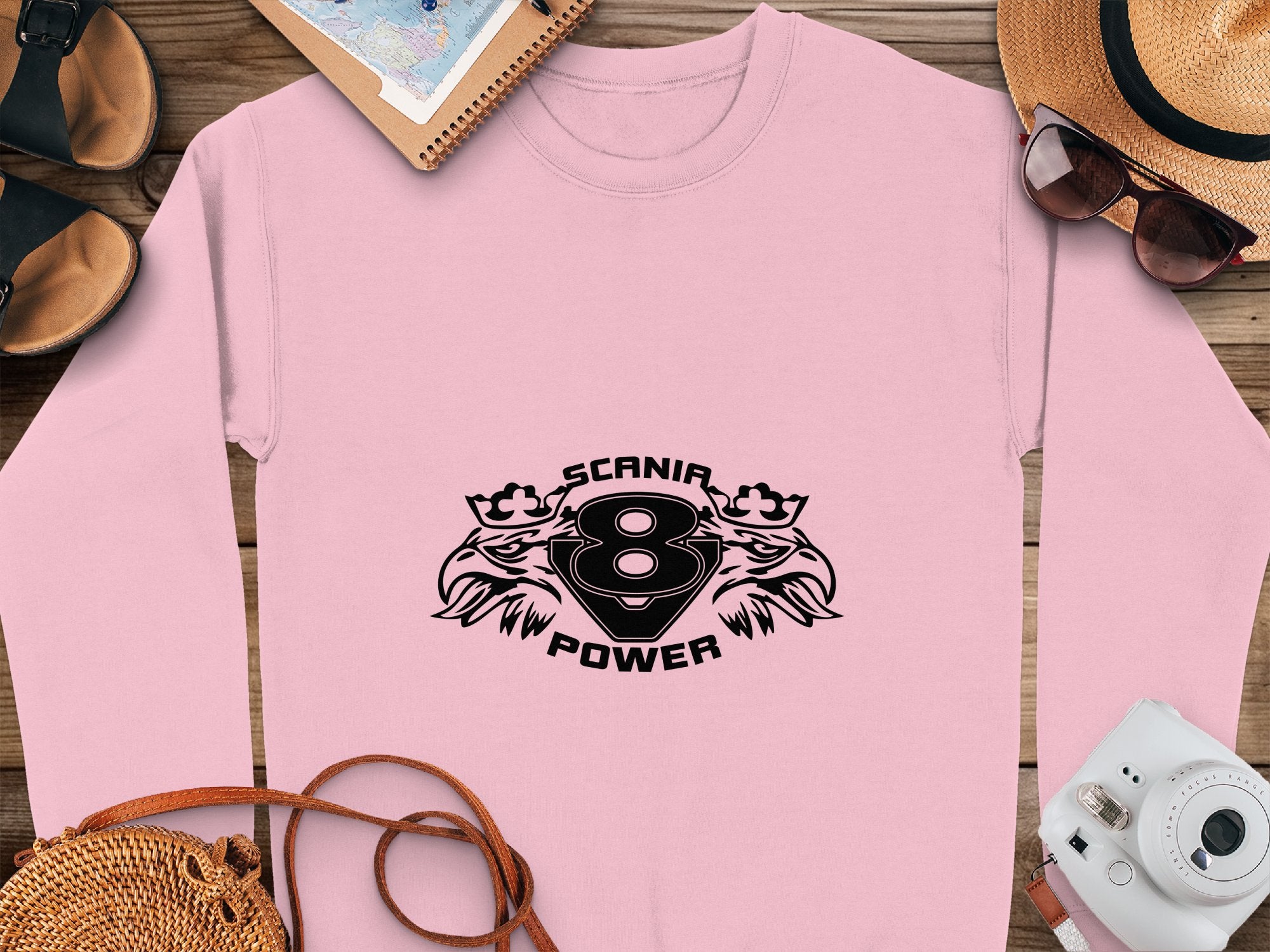 A trendy pink sweatshirt by Garment Graphics showcases SCANIR POWER, with two stylized eagle heads and the number 8. Paired with sandals, a notebook, sunglasses, a hat, and a camera on a wooden surface. Ideal for Scania Power V8 fans.
