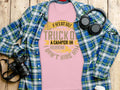 A Garment Graphics pink T-shirt featuring a humorous hiring quote is styled with a blue plaid shirt on rustic wood, accompanied by black headphones and a camera with strap—ideal for anyone who enjoys humor in their wardrobe essentials.