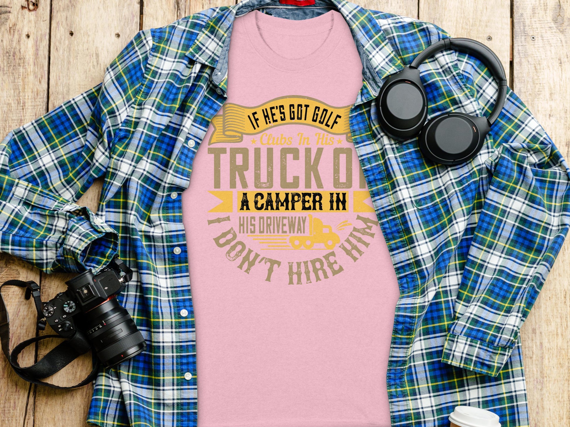 A Garment Graphics pink T-shirt featuring a humorous hiring quote is styled with a blue plaid shirt on rustic wood, accompanied by black headphones and a camera with strap—ideal for anyone who enjoys humor in their wardrobe essentials.
