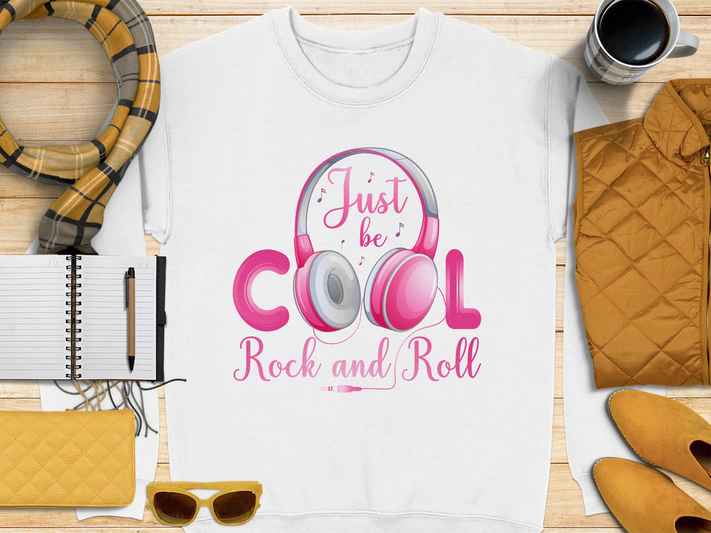 A white Garment Graphics shirt with pink headphones and Just be Cool Rock and Roll text is ideal for music enthusiasts. Styled alongside a plaid scarf, notebook, yellow sunglasses, purse, orange quilted jacket, and coffee cup on wood—a must-have for fashion lovers.