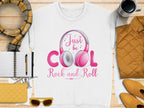 A white Garment Graphics shirt with pink headphones and Just be Cool Rock and Roll text is ideal for music enthusiasts. Styled alongside a plaid scarf, notebook, yellow sunglasses, purse, orange quilted jacket, and coffee cup on wood—a must-have for fashion lovers.