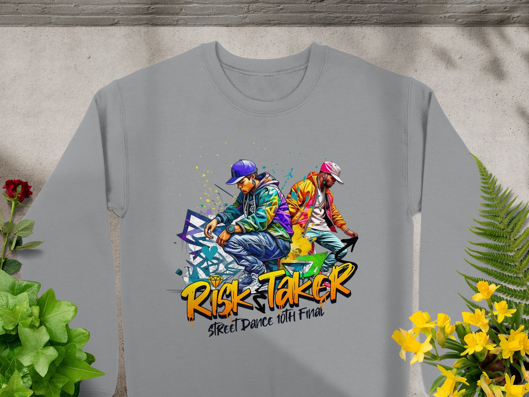This stylish gray sweatshirt by Garment Graphics showcases two street dancers in colorful outfits, with bold Risk Taker Street Dance 10th Final text. Laid on concrete, its surrounded by green and yellow plants, perfect for street dance enthusiasts.
