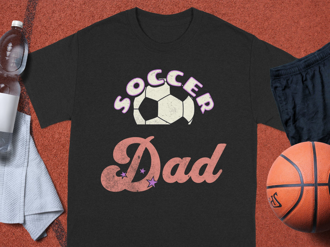 A Garment Graphics black T-shirt features Soccer Dad in stylized letters and a soccer ball graphic. Its displayed on a sports surface with a basketball, black shorts, water bottle, and white towel. Ideal for sporty dads balancing basketball on the court and soccer on the field.