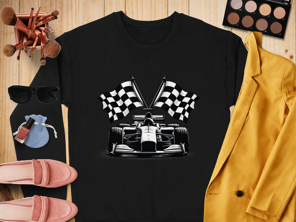 A flat lay showcases a black Garment Graphics sweatshirt with a racing car and checkered flag print, paired with a yellow blazer, sunglasses, makeup palette and brushes, nail polish, pink loafers, and a blue pouch on a wooden surface.