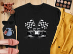 A flat lay showcases a black Garment Graphics sweatshirt with a racing car and checkered flag print, paired with a yellow blazer, sunglasses, makeup palette and brushes, nail polish, pink loafers, and a blue pouch on a wooden surface.