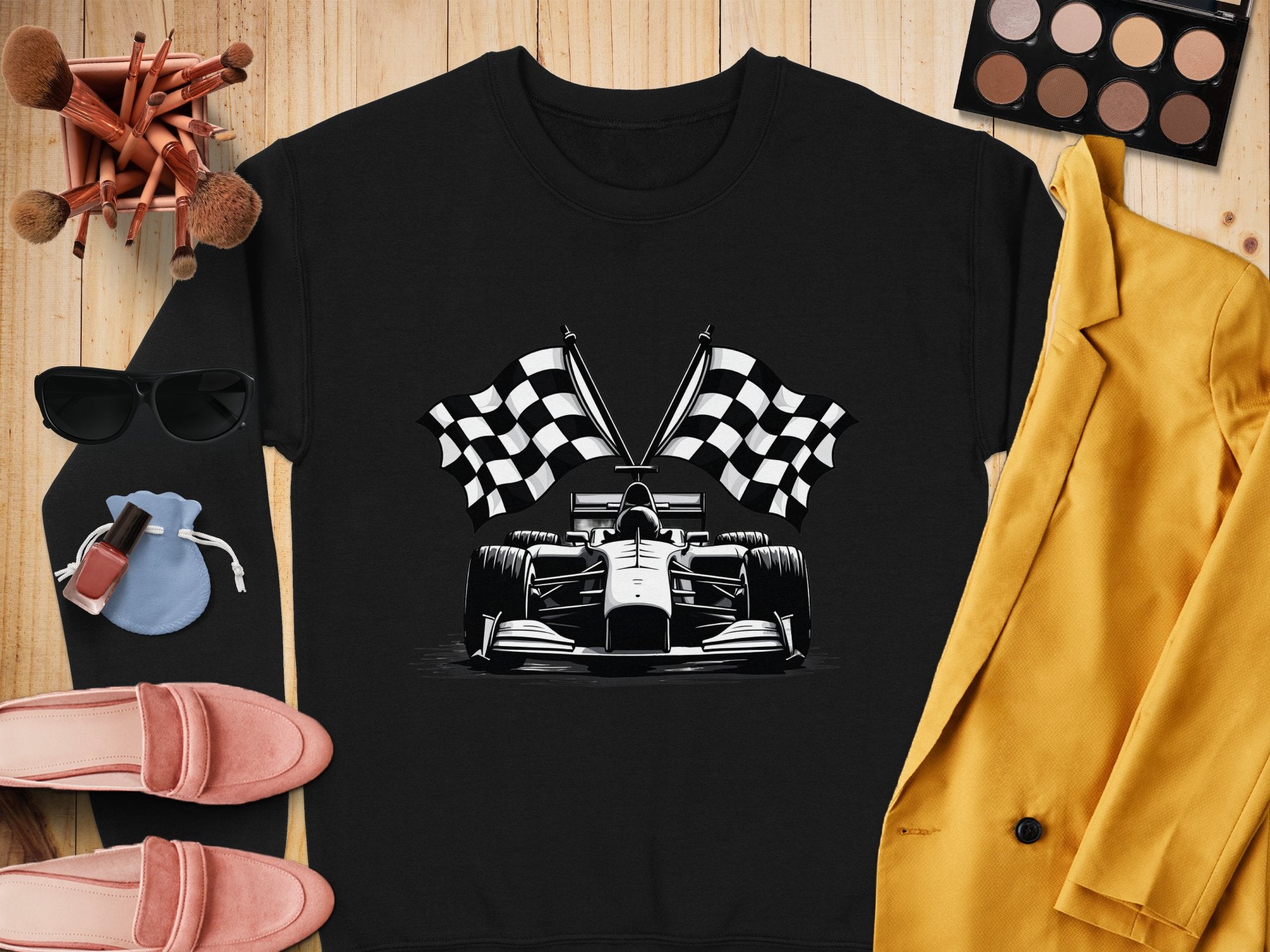 A flat lay showcases a black Garment Graphics sweatshirt with a racing car and checkered flag print, paired with a yellow blazer, sunglasses, makeup palette and brushes, nail polish, pink loafers, and a blue pouch on a wooden surface.