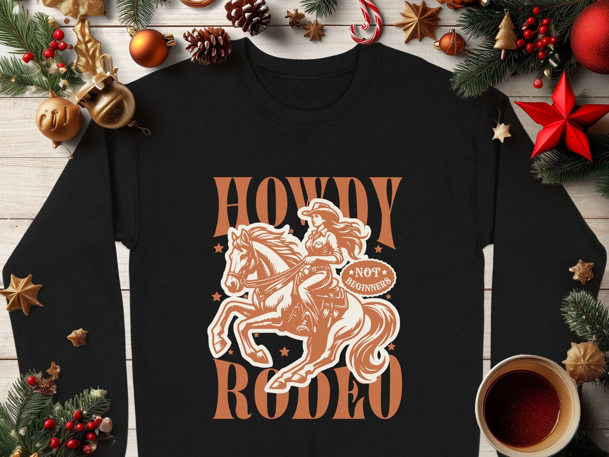 Garment Graphics black long-sleeve shirt features a cowgirl and horse illustration with Howdy Rodeo text. Its set on wood, surrounded by Christmas decor and coffee, making it a perfect gift for rodeo fans.