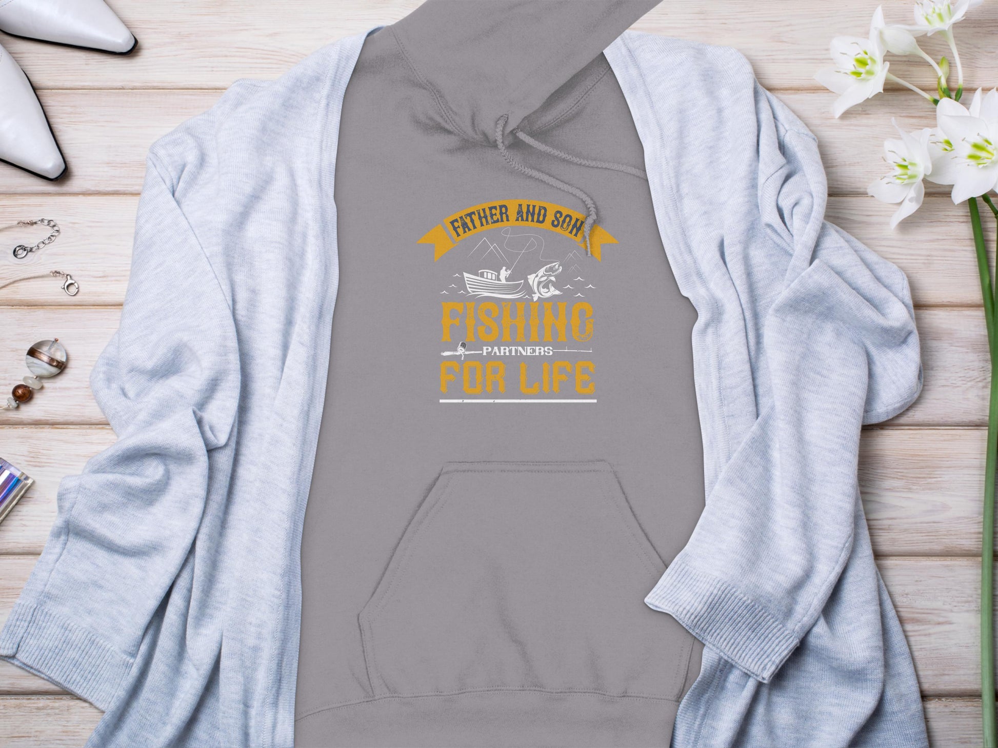 Garment Graphics gray fishing hoodie features Father and Son Fishing Partners for Life printed in yellow and white. Displayed on a wooden surface with a light blue cardigan, white flowers, silver jewelry, a watch, and white shoes—ideal for fishing enthusiasts celebrating their bond.