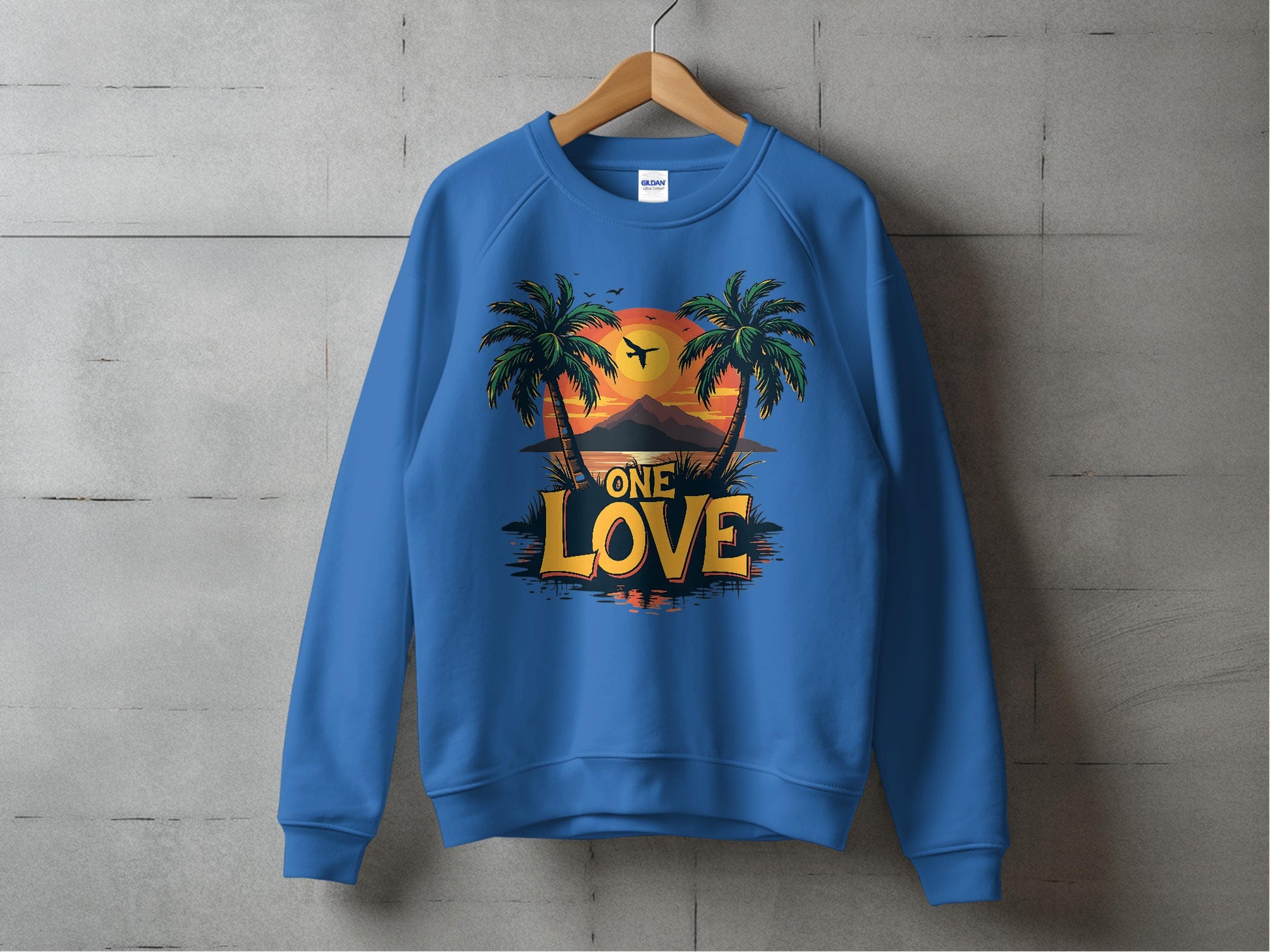 A blue sweatshirt by Garment Graphics hangs on a wooden hanger against a concrete wall, featuring an eye-catching design of palm trees and a vibrant sunset. Beneath the scenic artwork, the words ONE LOVE stand out boldly.