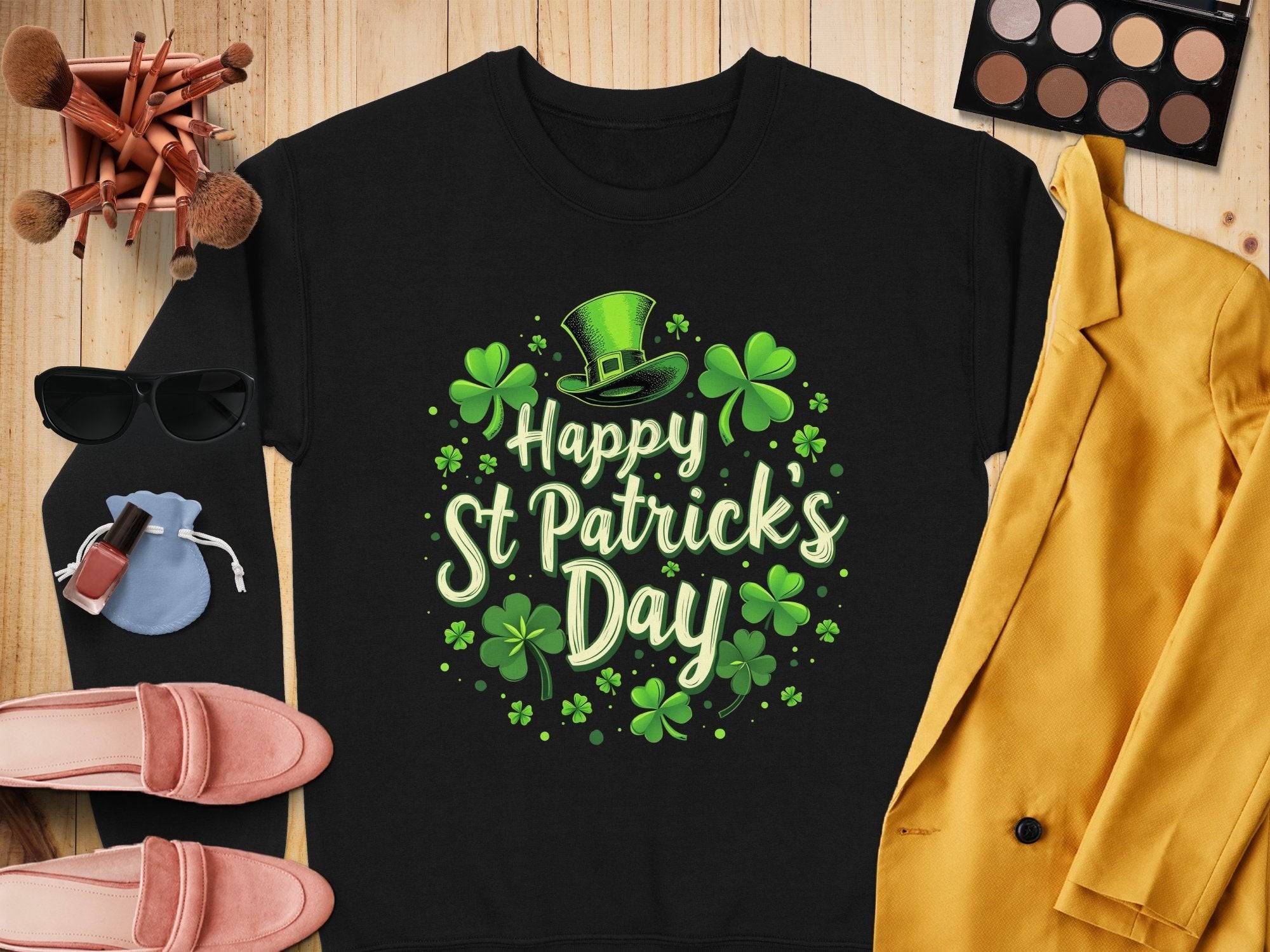 Flat lay of a black Garment Graphics sweatshirt with shamrocks and a leprechaun hat for St. Patricks Day, surrounded by a yellow jacket, pink shoes, sunglasses, makeup brushes, and an eyeshadow palette on wood.