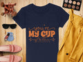 A stylish flat lay features a Garment Graphics 100% cotton navy t-shirt with the orange Youre My Cup of Tea graphic. It is elegantly arranged with a yellow blazer, pink loafers, sunglasses, makeup palette, brushes, watch, and nail polish on a wooden surface.