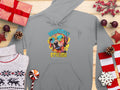 A Garment Graphics gray hoodie displays a playful dog in blue sunglasses with the text Pawsitively Stylish, set amidst holiday-themed items like a sweater, wrapped gifts, pinecones, and decorations on a wooden surface.