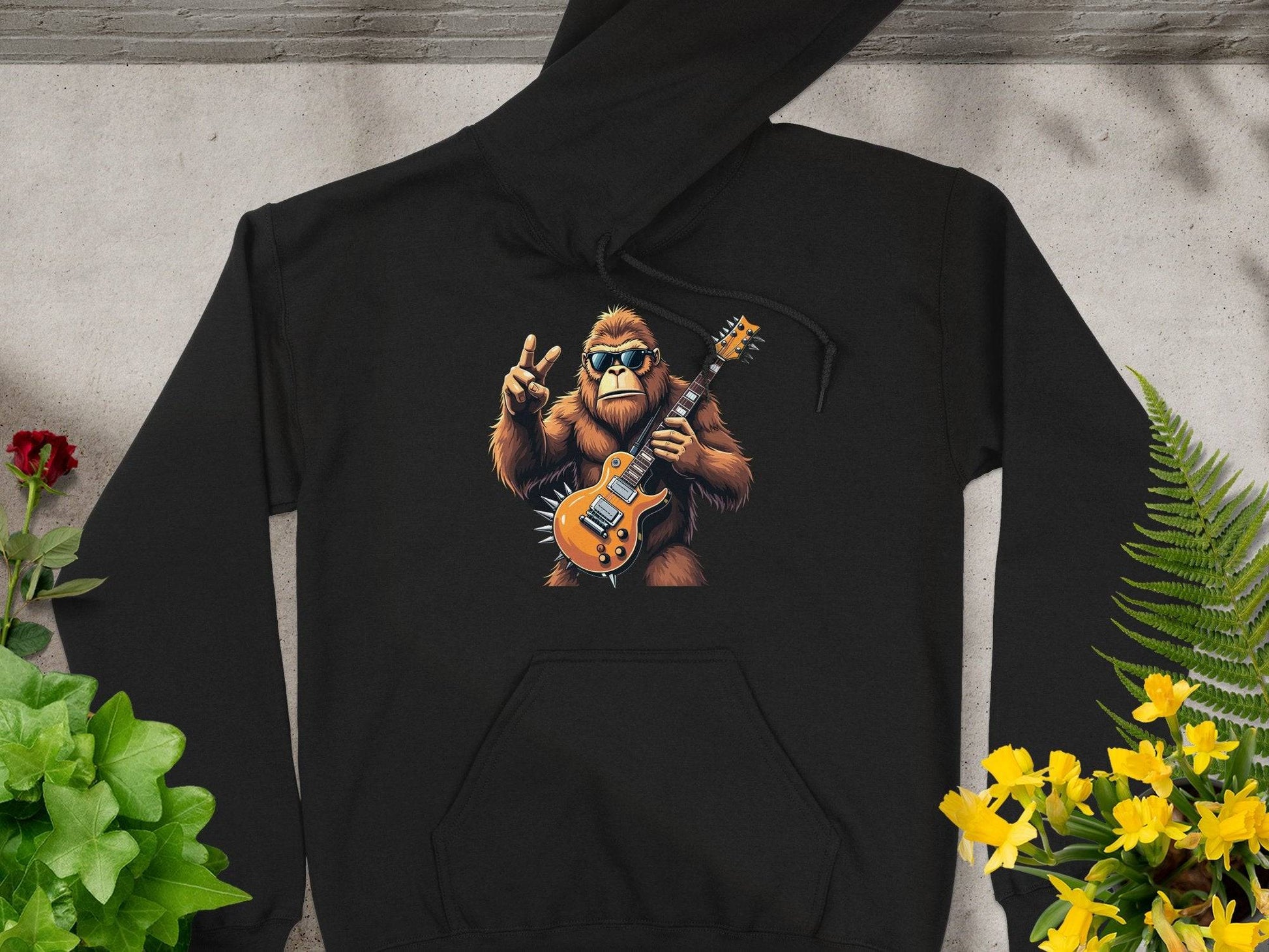 The Garment Graphics black gorilla hoodie features a cool cartoon gorilla sporting sunglasses and playing an electric guitar with a peace sign. Set on a gray surface with flowers, its ideal for music enthusiasts.