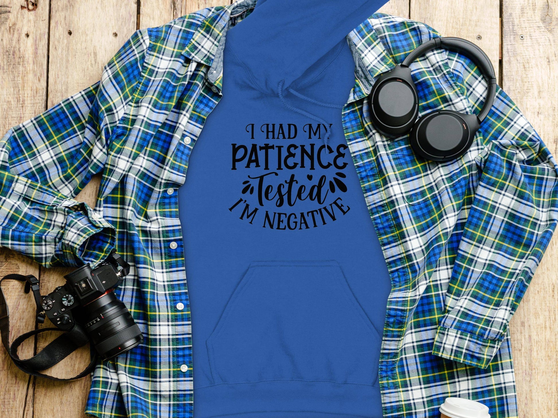 The Garment Graphics blue hoodie features black text saying, I had my patience tested Im negative. Laid under a blue/green plaid shirt, it boasts a medium-heavy fabric and classic fit. Nearby are a camera, black headphones, and a blue mug on a wooden surface.