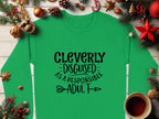 A green Garment Graphics shirt with Cleverly disguised as a responsible adult in black font is centered on a wooden surface, surrounded by festive pine branches, ornaments, and a red bow. A cup of coffee sits at the bottom right. Perfect for your humor wardrobe and unisex sizing needs.