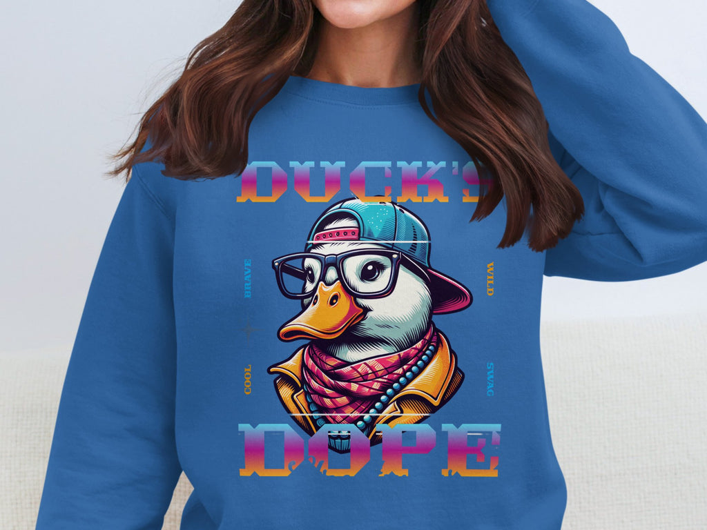 Wearing a stylish blue Garment Graphics sweatshirt, featuring a quirky duck with glasses, cap, and scarf. The bold fashion statement reads DUCKS DOPE, surrounded by words like COOL, BRAVE, WILD, and SPACE for a playful yet edgy vibe.
