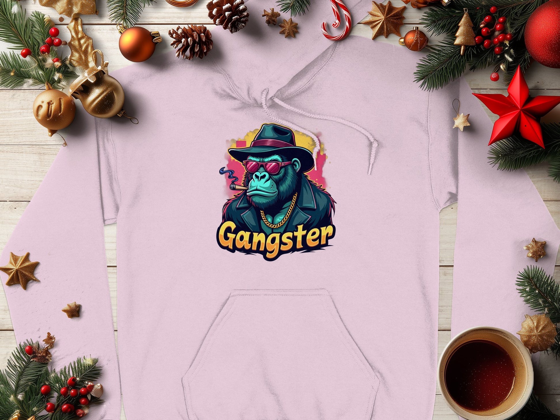 The Garment Graphics light pink graphic hoodie showcases a gangster ape with a hat, sunglasses, and a cigarette. Gangster is boldly printed underneath. This trendy piece is set among Christmas decor and a steaming coffee cup on wood.