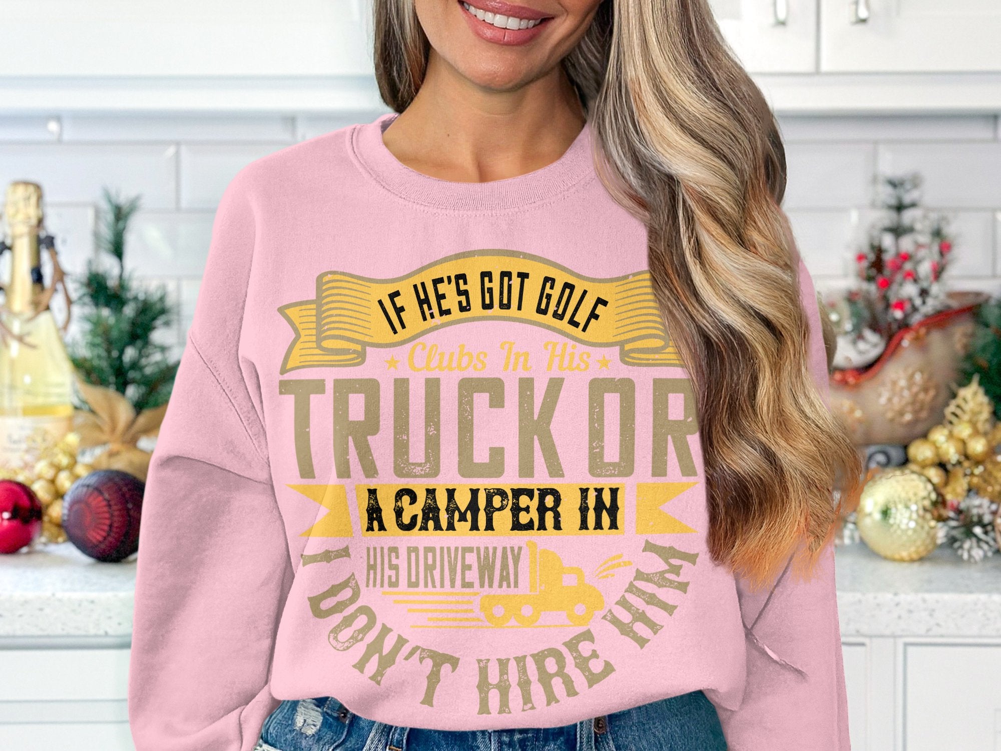 A person in a humorous pink Garment Graphics sweatshirt that reads If hes got golf clubs in his truck or a camper in his driveway, dont hire him. stands in a kitchen decorated with holiday ornaments and small trees.