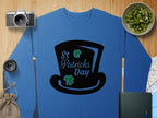 Garment Graphics blue sweatshirt, showcasing a black top hat labeled St Patricks Day with two green shamrocks, exudes festive spirit. It is set on a wooden surface surrounded by a camera, lens, map, bowl of kiwi, green book, and burlap sack.