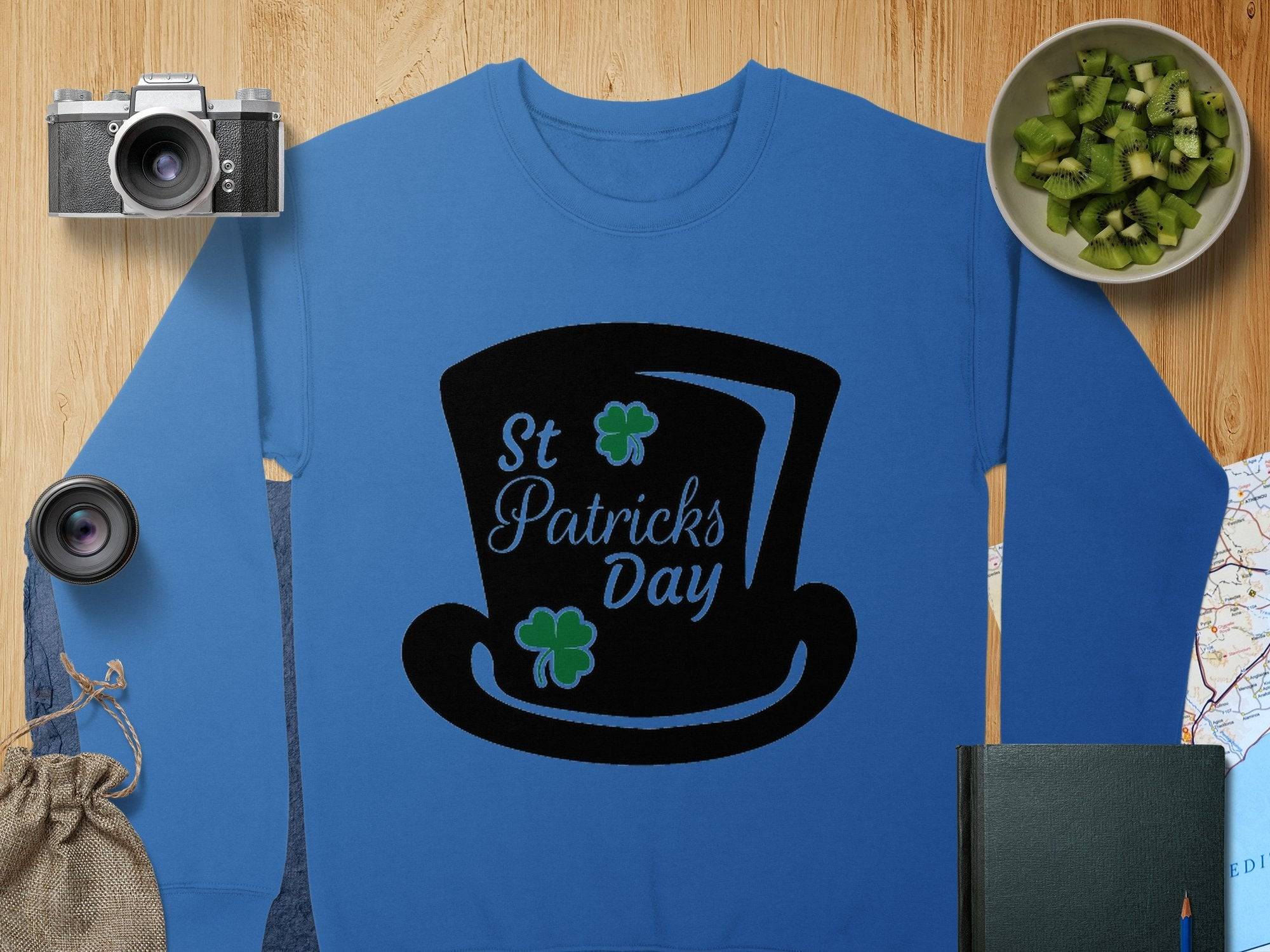 Garment Graphics blue sweatshirt, showcasing a black top hat labeled St Patricks Day with two green shamrocks, exudes festive spirit. It is set on a wooden surface surrounded by a camera, lens, map, bowl of kiwi, green book, and burlap sack.