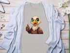 A Garment Graphics gray eagle T-shirt, with a cartoon eagle giving a thumbs-up, rests on a light wooden surface. Its partially covered by a light blue cardigan and surrounded by white flowers and jewelry, enhancing its fun, classic fit appeal.