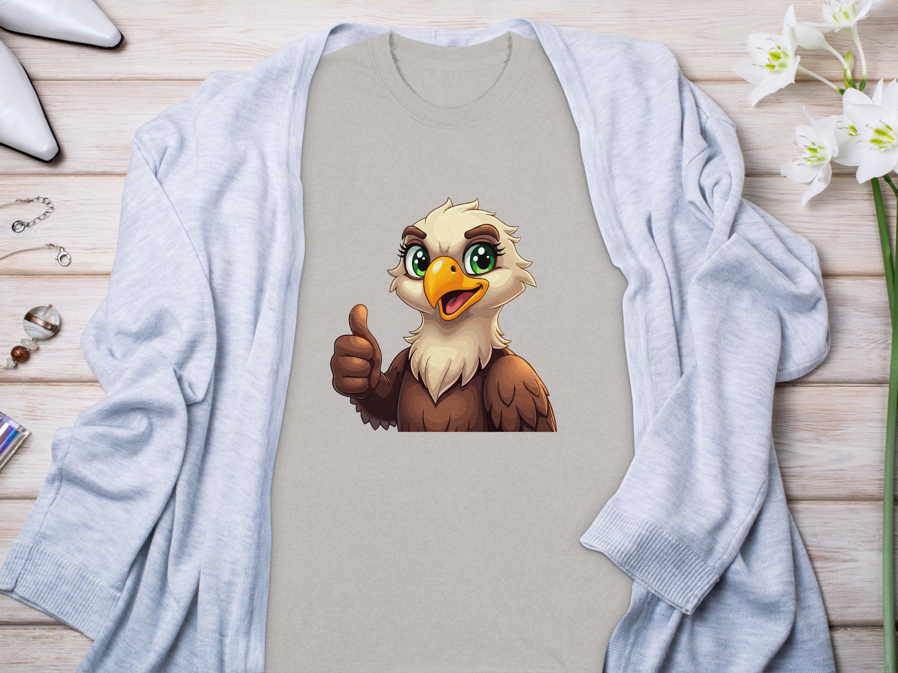 A Garment Graphics gray eagle T-shirt, with a cartoon eagle giving a thumbs-up, rests on a light wooden surface. Its partially covered by a light blue cardigan and surrounded by white flowers and jewelry, enhancing its fun, classic fit appeal.