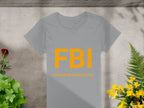 A Garment Graphics gray FBI Female Boob Inspector T-Shirt, featuring FBI in bold yellow letters with FEMALE BOOB INSPECTOR below, is laid on a concrete surface surrounded by plants. Ideal for any occasion as a gag gift.
