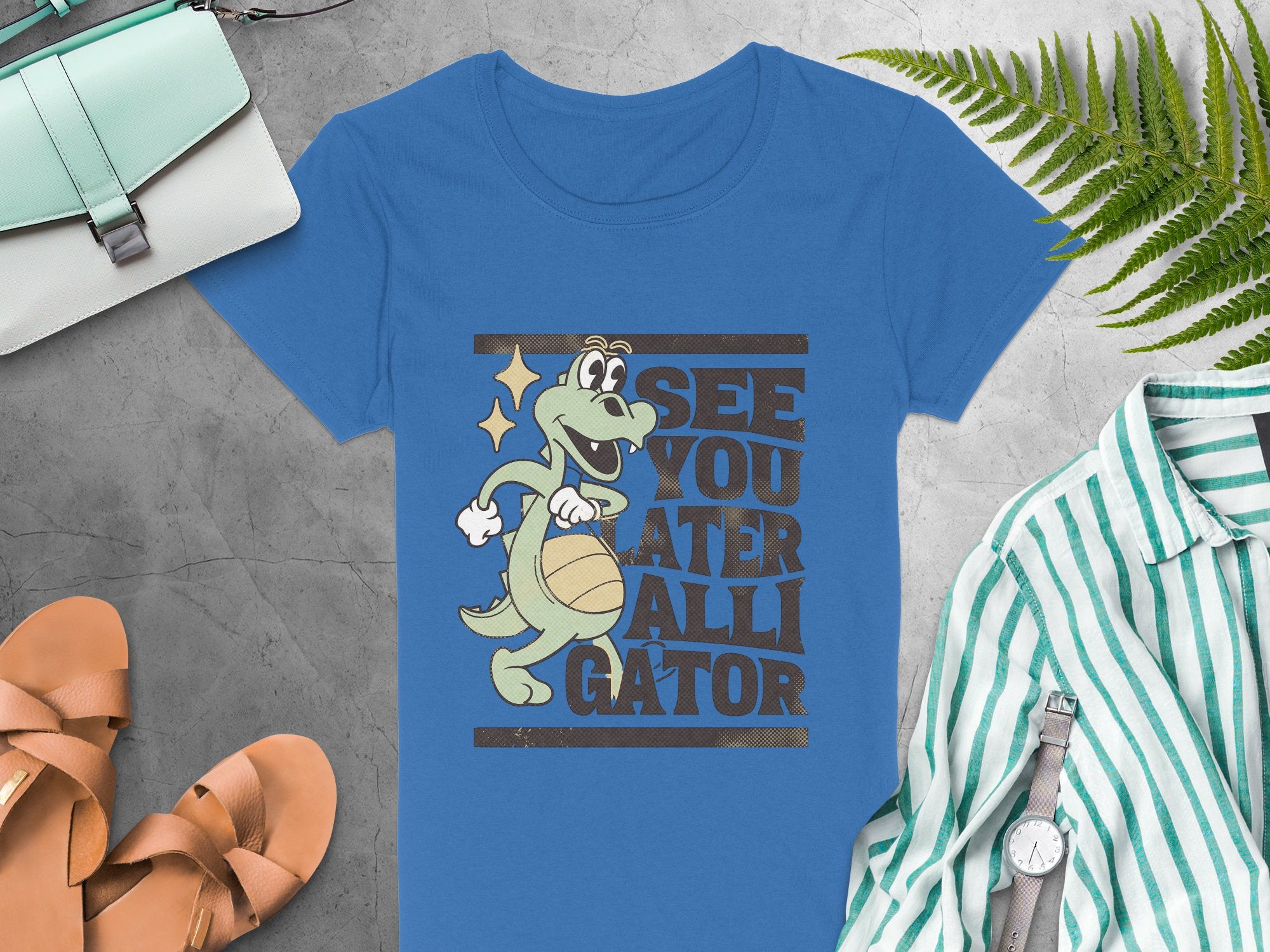 Garment Graphics presents a blue womens alligator t-shirt illustrating a smiling gator with a basketball and playful text See you later, alligator, displayed on gray with a purse, striped shirt, sandals, and fern leaf.
