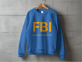 Displayed on a gray concrete wall, this Garment Graphics unisex graphic print sweatshirt features bold yellow FBI letters and the witty Female Boob Inspector text on blue fabric hanging from a wooden hanger.