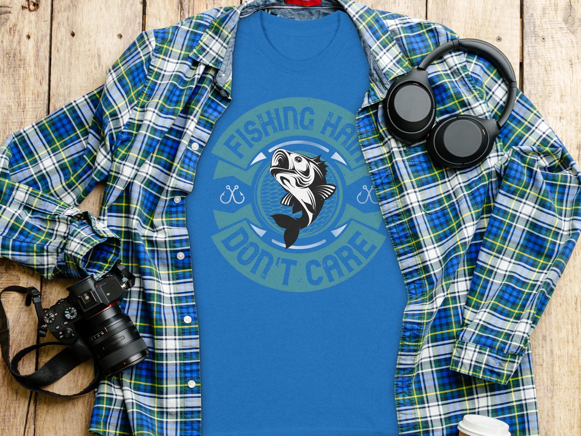 A flat lay features a blue Garment Graphics tee with Fishing Hair Dont Care and a fish design, surrounded by a plaid shirt, distinctive headphones, a camera, and a white travel mug on wood.