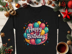 Garment Graphics offers a black long-sleeve unisex shirt with colorful balloons and Happy Birthday text, ideal for celebrations. The shirt pairs beautifully with festive decorations like pine branches, ornaments, and a cozy cup of coffee on a wooden surface.