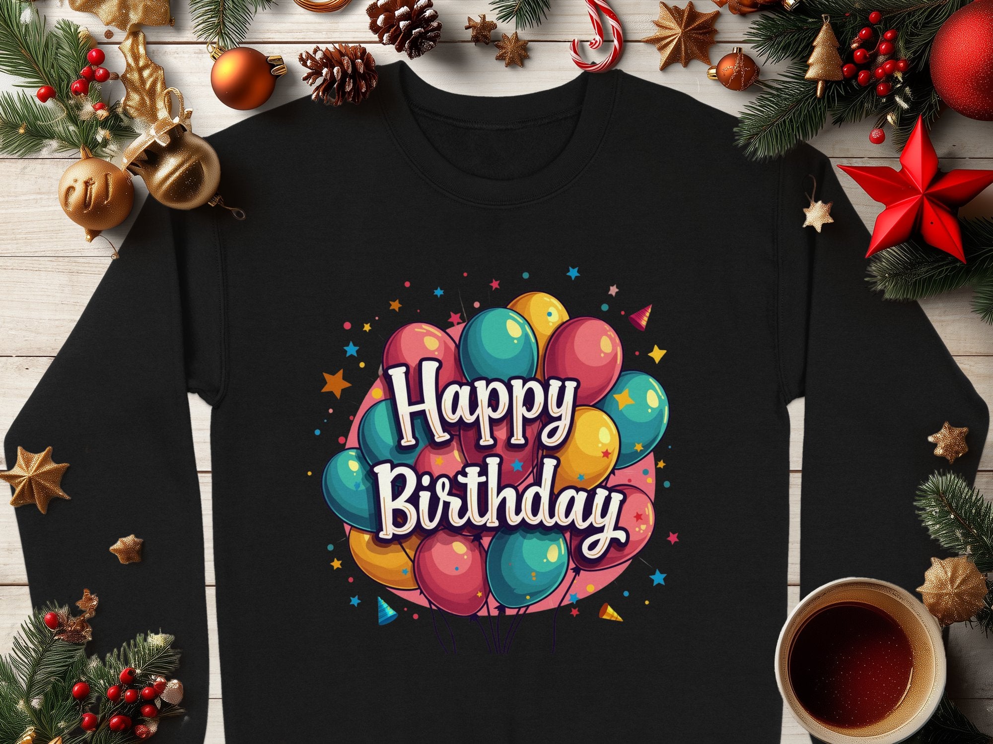 Garment Graphics offers a black long-sleeve unisex shirt with colorful balloons and Happy Birthday text, ideal for celebrations. The shirt pairs beautifully with festive decorations like pine branches, ornaments, and a cozy cup of coffee on a wooden surface.