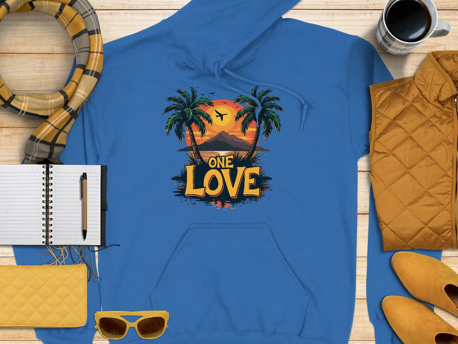 Garment Graphics blue graphic hoodie, adorned with a One Love motif depicting a tropical sunset with palm trees and a bird in vivid colors, sits on a rustic wooden surface alongside nearby items: a yellow quilted purse, sunglasses, scarf, jacket, notebook with pen, and coffee mug.