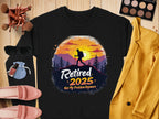 Flat lay showcasing Garment Graphics black unisex t-shirt featuring a hiker silhouette against a sunset with Retired 2025 Not My Problem Anymore, surrounded by sunglasses, makeup, pink loafers, and a mustard blazer on a wooden surface.