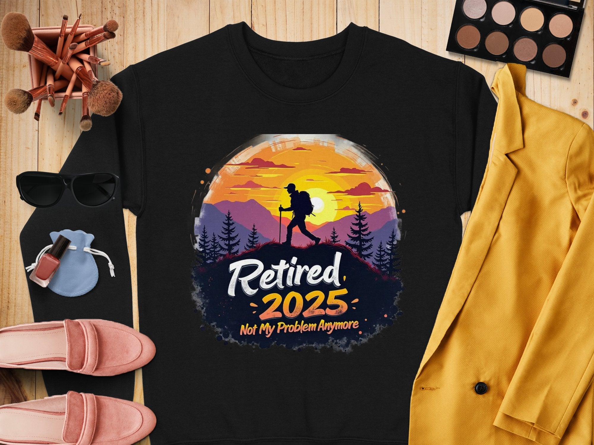 Flat lay showcasing Garment Graphics black unisex t-shirt featuring a hiker silhouette against a sunset with Retired 2025 Not My Problem Anymore, surrounded by sunglasses, makeup, pink loafers, and a mustard blazer on a wooden surface.
