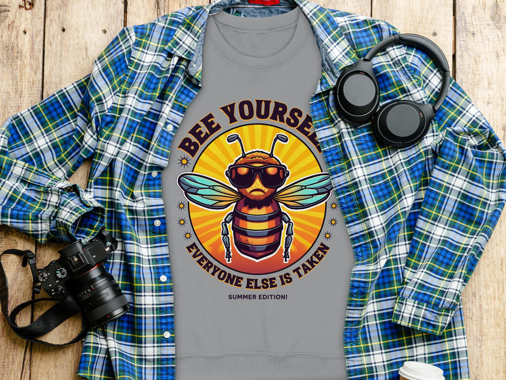 The Garment Graphics unisex T-shirt features a bee with sunglasses and headphones, alongside the text Bee Yourself, Everyone Else is Taken, Summer Edition, displayed on a plaid shirt next to a camera, headphones, and a bottle on wood.