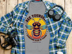 The Garment Graphics unisex T-shirt features a bee with sunglasses and headphones, alongside the text Bee Yourself, Everyone Else is Taken, Summer Edition, displayed on a plaid shirt next to a camera, headphones, and a bottle on wood.