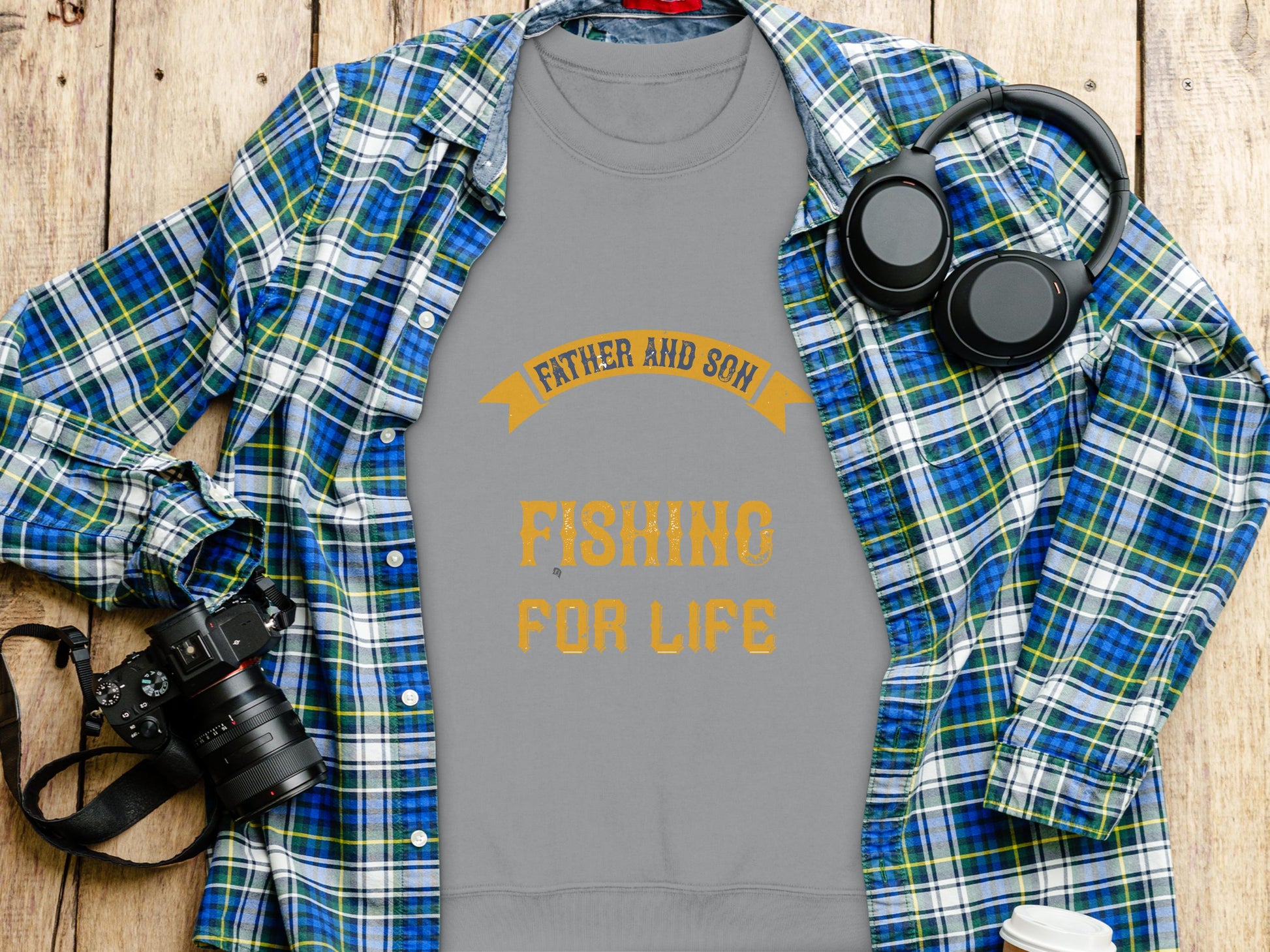 The grey Garment Graphics sweatshirt showcases Father and Son Fishing for Life in vibrant orange text, resting atop a blue plaid shirt and accompanied by headphones, a camera, and a white coffee cup on a wooden surface.